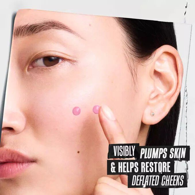 Model points to two pink serum drops on cheek beside text reading “Visibly plumps skin & help restore depleted cheeks,” highlighting a skincare product’s plumping benefit.​