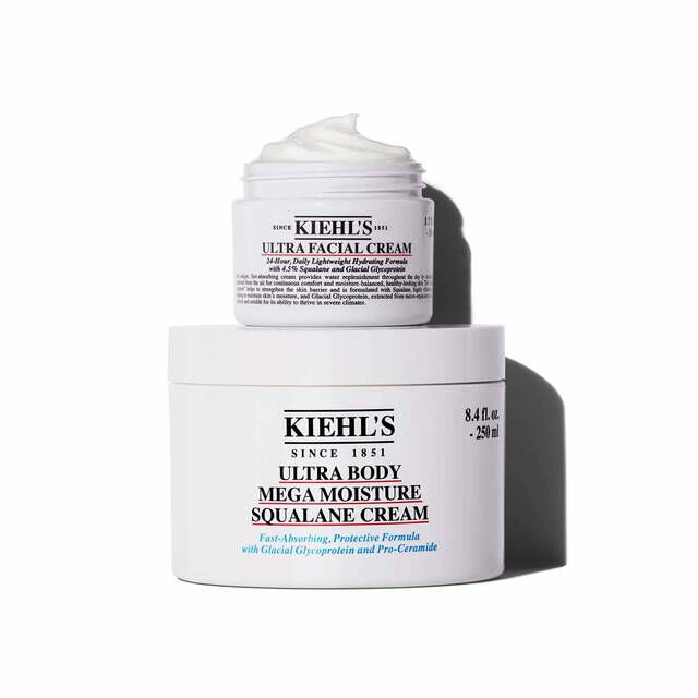 A graphic featuring two white jars of Kiehl’s cream, stacked one on top of the other, casting a shadow against a white background. The smaller top jar is "KIEHL’S ULTRA FACIAL CREAM" with a visible swirl of white cream. The larger bottom jar is "KIEHL’S ULTRA BODY MEGA MOISTURE SQUALANE CREAM."