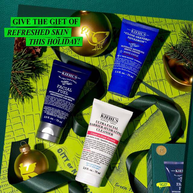 A festive graphic, set against a green background with a map pattern, features three Kiehl's products: a dark blue tube of "KIEHL’S FACIAL MOISTURUZER," a dark blue tube of "KIEHL’S CLOSE-SHAVERS SQUADRON ULTIMATE BRUSHLESS SHAVE CREAM," and a white tube of "KIEHL’S ULTRA FACIAL BARRIER-HYDRATING CLEANSER." Two gold Christmas ornaments, one with a yellow bird design and another with a yellow "K" logo, are part of the arrangement, along with green ribbon and pine branches. A partial view of a green Kiehl's gift box is visible in the lower right. Bright green text in the upper left reads "GIVE THE GIFT OF REFRESHED SKIN THIS HOLIDAY!"