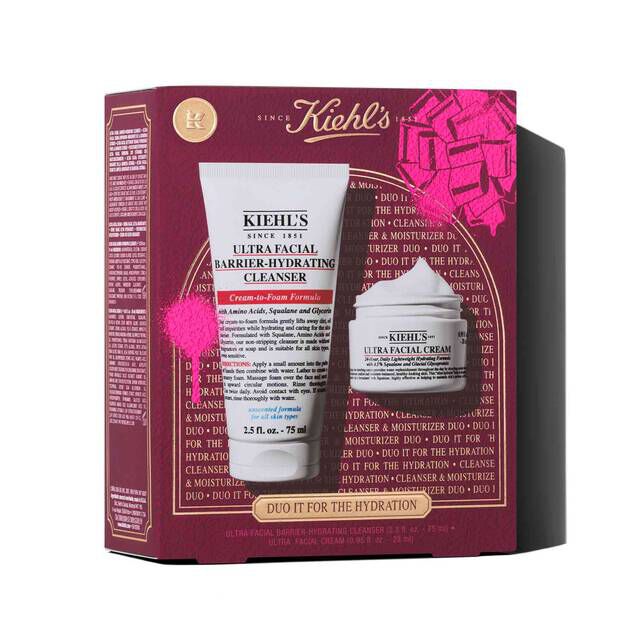 A graphic displays a Kiehl's gift set box, in dark red, with a pink bow graphic in the upper right corner and a pink paint splatter effect in the lower left. The box states "DUO IT FOR THE HYDRATION" on its front and features two products: a white tube of "KIEHL'S ULTRA FACIAL BARRIER-HYDRATING CLEANSER" and a white jar of "KIEHL'S ULTRA FACIAL CREAM".