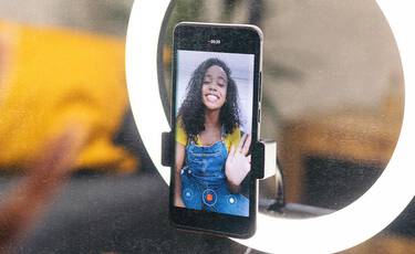 Smartphone mounted in a ring light showing a young woman waving on screen, depicting digital content creation.​