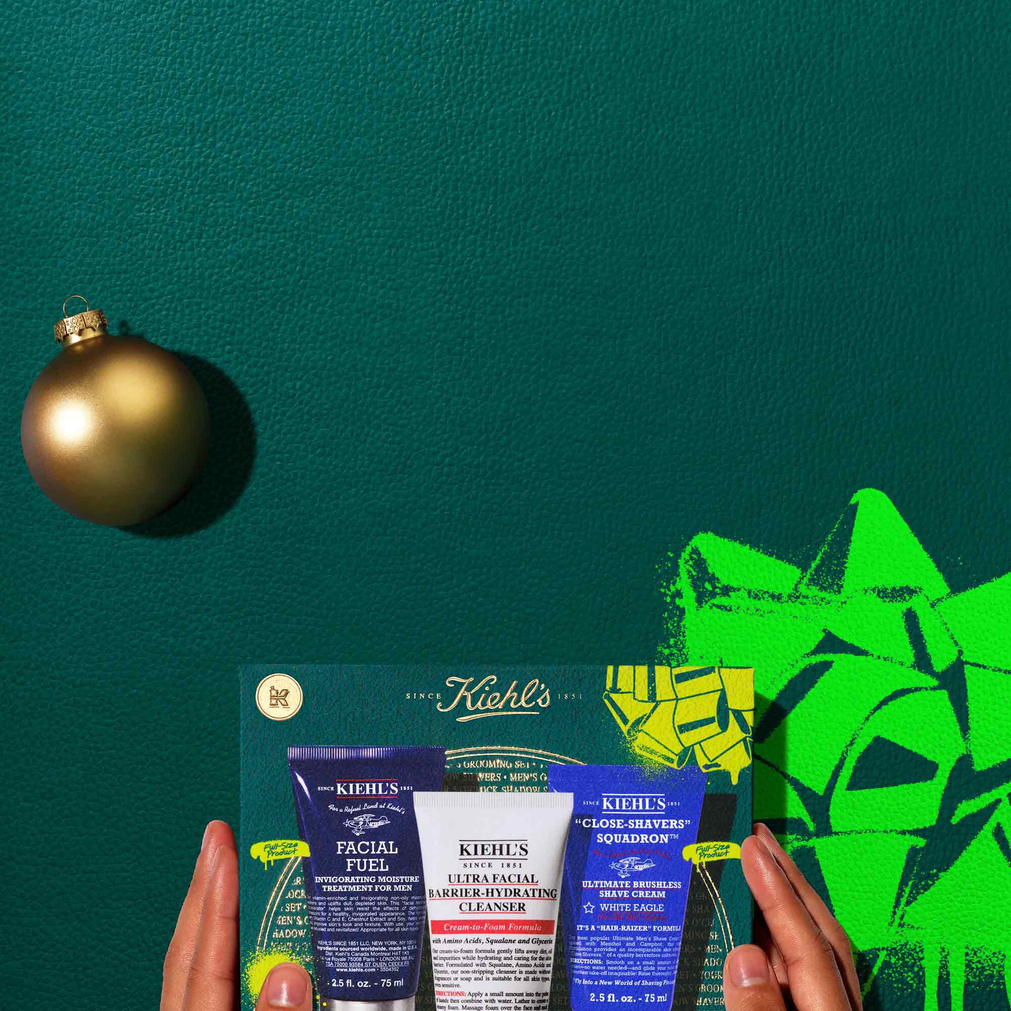 A graphic, set against a dark green textured background, features three Kiehl’s products and a gift box. The top row displays a dark blue tube of "KIEHL’S FACIAL FUEL ENERGIZING MOISTURIZER," a white tube of "KIEHL’S ULTRA FACIAL BARRIER-HYDRATING CLEANSER," and a dark blue tube of "KIEHL’S CLOSE-SHAVERS SQUADRON ULTIMATE BRUSHLESS SHAVE CREAM." A gold spherical Christmas ornament is also present on the left. Below these, a dark teal Kiehl’s gift box showcases the same three products, decorated with bright green graphic paint splatters and a bow design.