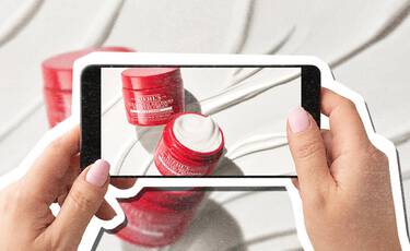 Smartphone photographing red Kiehl’s Super Multi- Corrective Cream jars against a white textured background.​